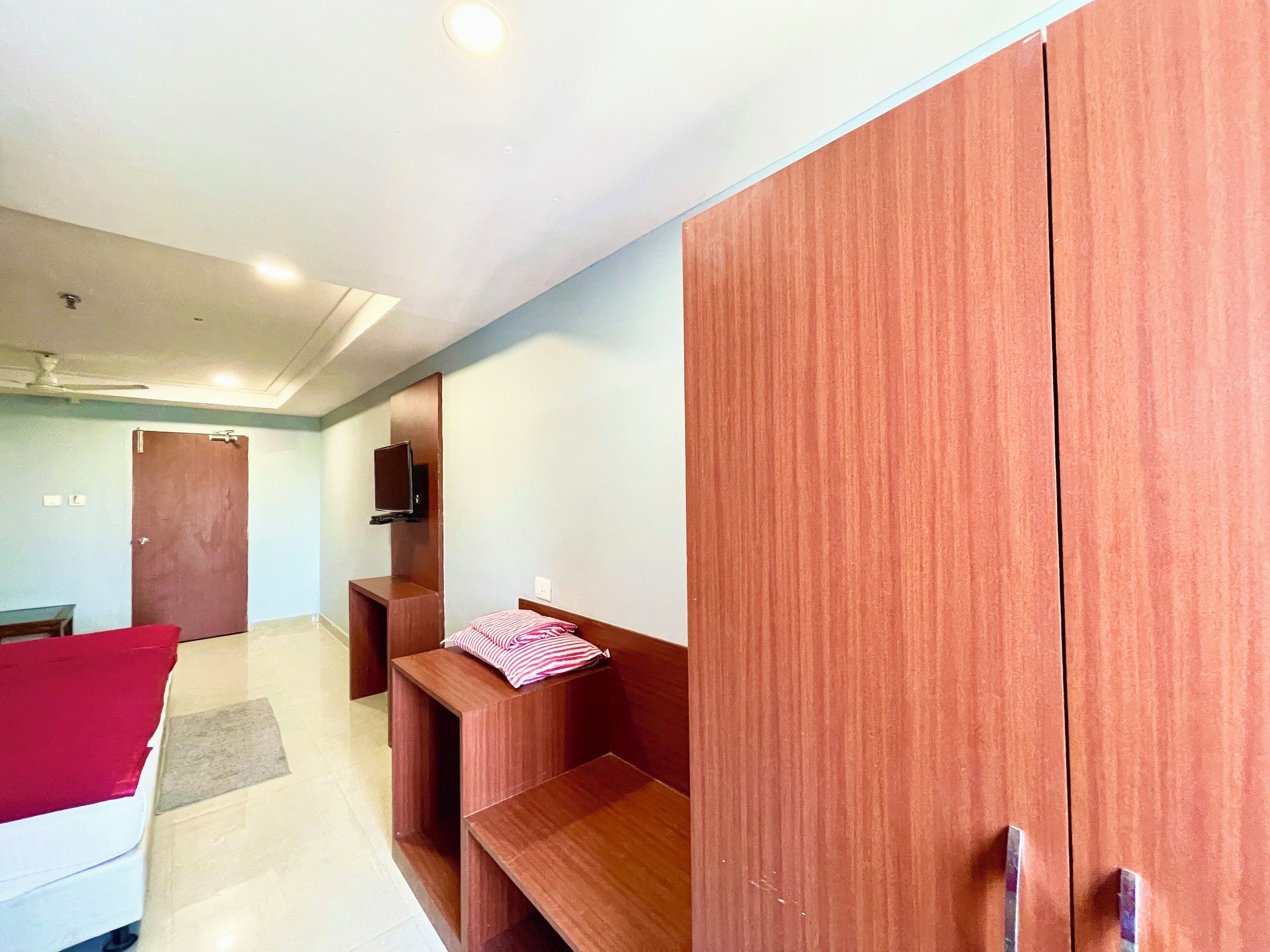 A hotel room with a bed, a wooden wardrobe, a TV mounted on the wall, and a ceiling fan.