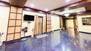 A spacious lobby with a marble floor, a flat-screen TV mounted on the wall, a fan, and potted plants. - Hotel Janaki Pride - near Temple (Puri)