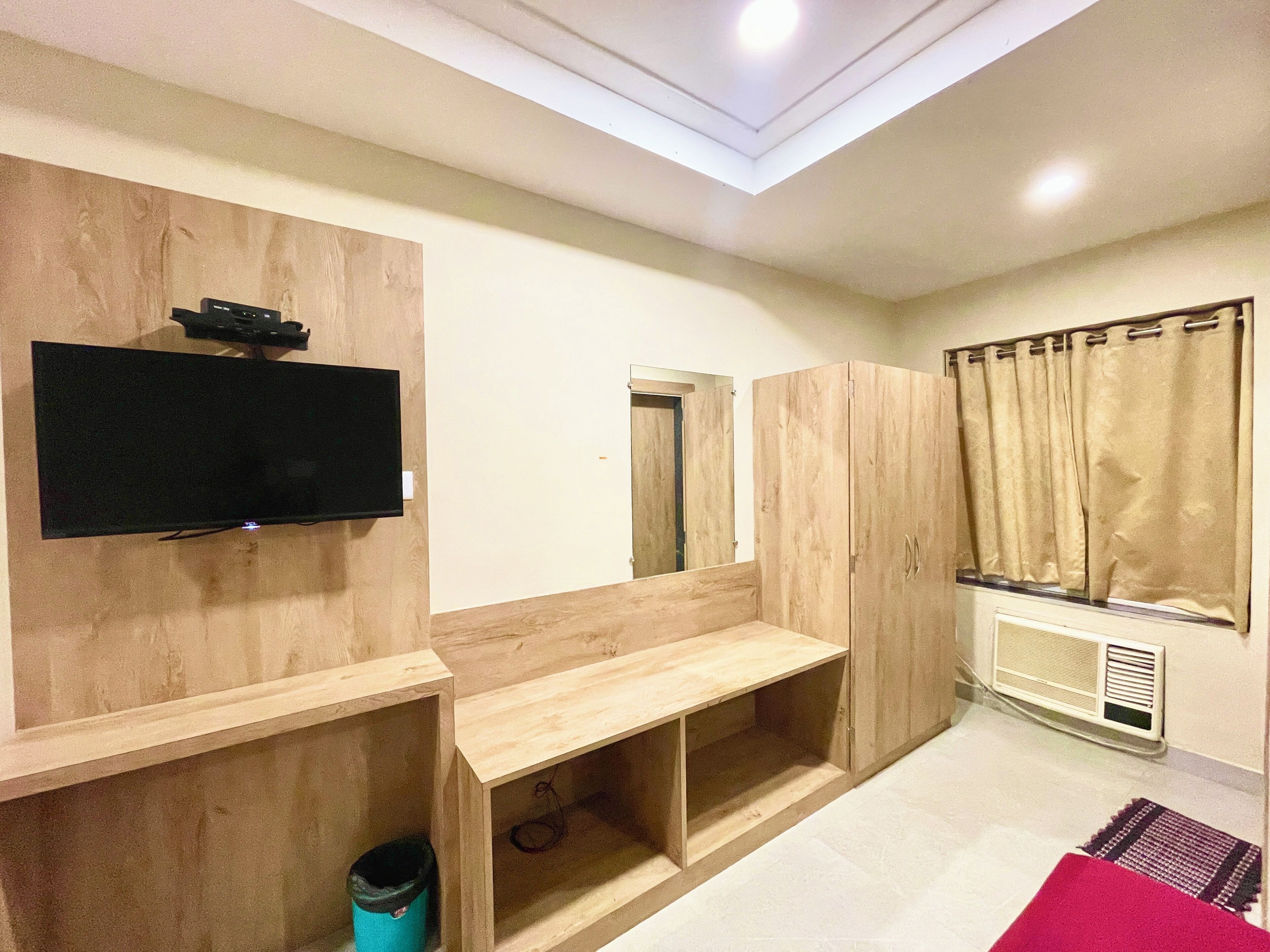 A room with a flat-screen TV mounted on a wooden wall, a wooden bench, a mirror, and a window with curtains.