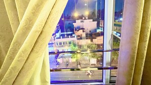 A view through a window with curtains, revealing a street scene with buildings and streetlights. - Hotel Janaki Pride - near Temple (Puri)
