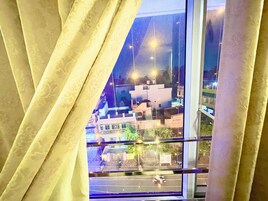 Une vue à travers une fenêtre avec des rideaux, dévoilant une scène de rue avec des bâtiments et des lampadaires.