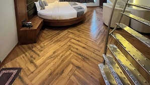 A modern hotel room with a circular bed, wooden flooring, a built-in bench, a desk, a mirror, a ceiling fan, a staircase, and a window with curtains. - The Saroja Grand Villa Resorts (Vasai)