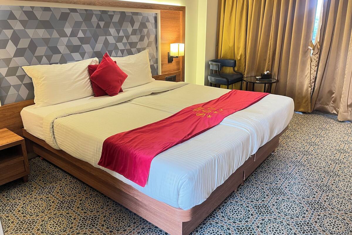 A hotel room with a large bed, a headboard with a geometric pattern, a red throw blanket, and a tiled floor.