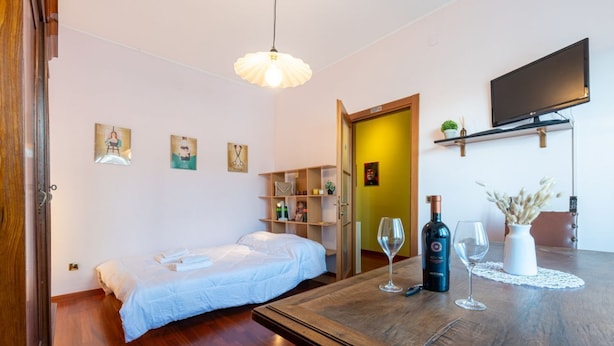 A room with a bed, a wooden table with a bottle and glasses, a TV, and shelves with various items. - Cosy apartment of 34 sqm, for 2 people, on the fourth floor of a building with lift (with some steps to access it).The property is located in a lively district and well served by public transport; the apartment is 50 m from the Hospital of Parma and a fe (Parma)