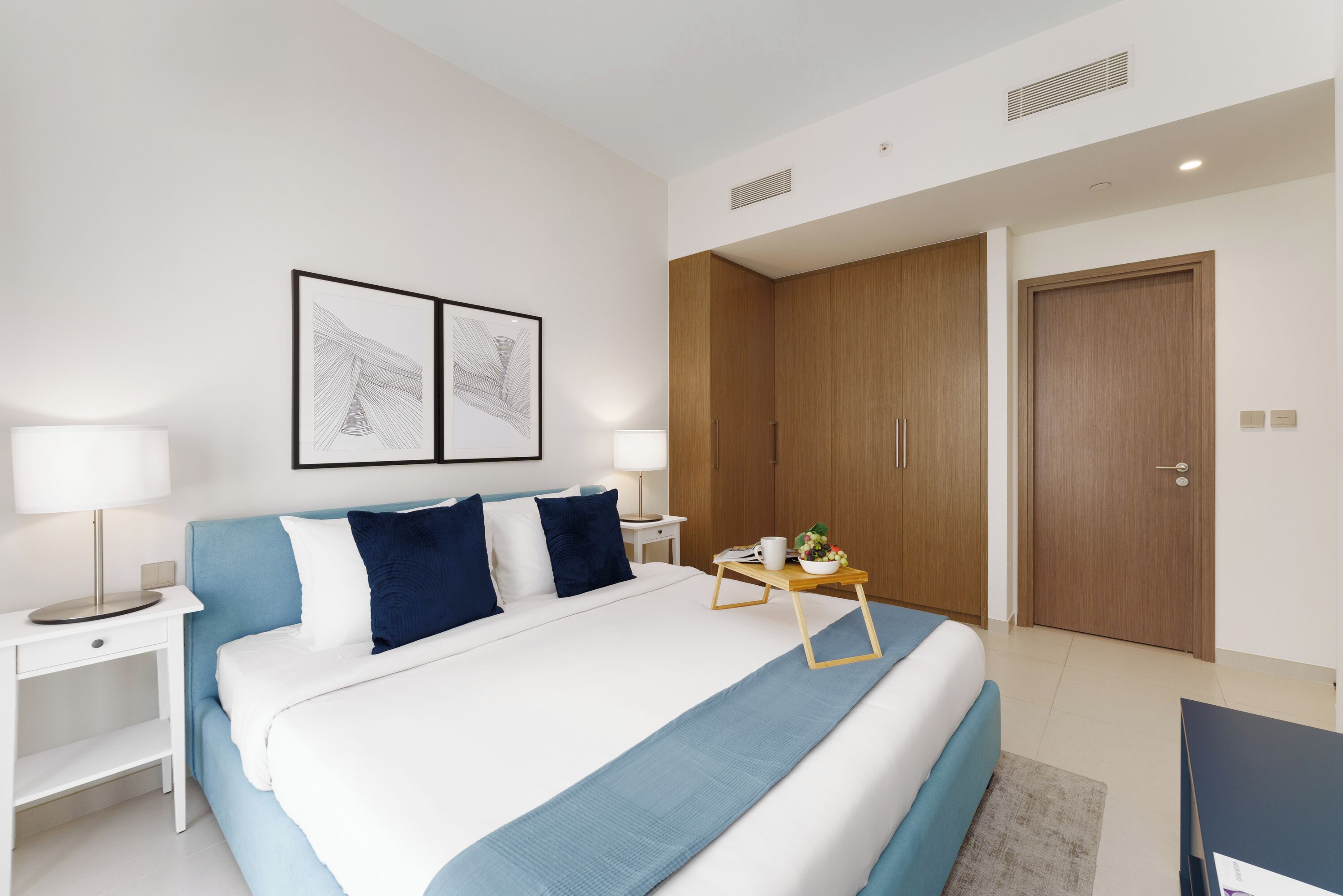 Una habitación de hotel moderna con una cama grande, una mesita de noche con lámpara, una pequeña mesa con un florero y un armario.