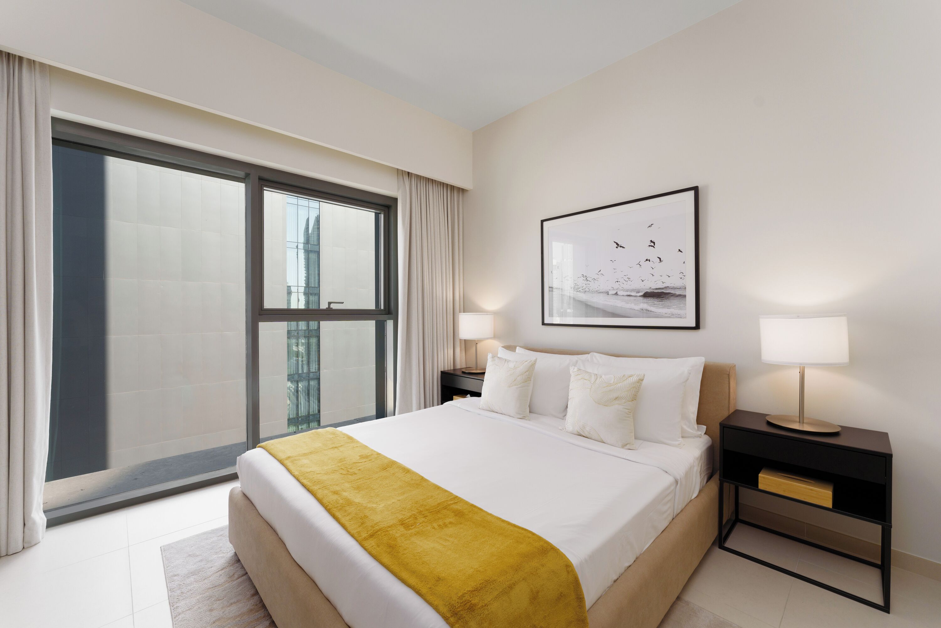 A modern hotel room with a large bed, a nightstand with a lamp, a framed picture on the wall, and a view of the city through the window.