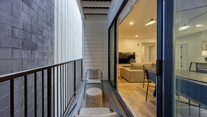 Un balcon étroit doté d’une rambarde en métal, de deux chaises, d’une petite table ronde et d’une porte vitrée donnant sur un espace intérieur comprenant un canapé, un téléviseur et un coin repas.