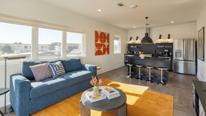 A modern living room with a blue sofa, a round coffee table with books and a vase, and a kitchen area with a bar and stainless steel appliances.
