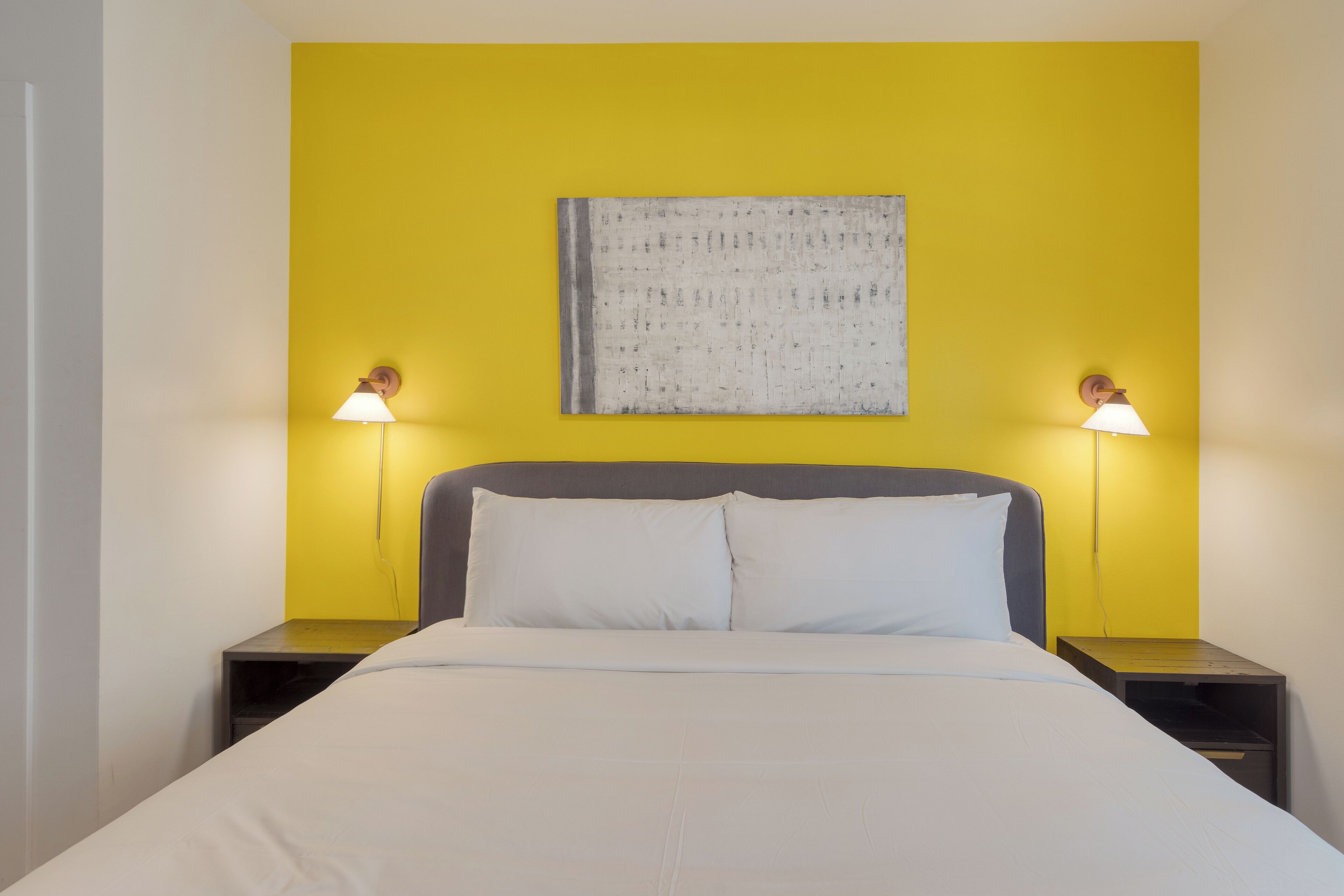 A bed with white bedding, two bedside tables with lamps, and a yellow wall with a textured artwork above the bed.