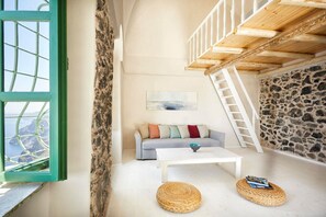 A cozy room with a stone wall, a white sofa with colorful cushions, a white table, and a view through a green-framed window.