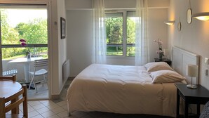 A bedroom with a bed, a bedside table with a lamp, a window with curtains, a chair, and a table with a potted plant. - Appartement hotel (Castera-Verduzan)