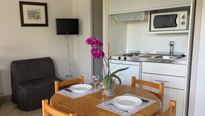 A dining area with a wooden table set for two, a dark armchair, a flat-screen TV mounted on the wall, and a kitchenette with a microwave and stove. - Appartement hotel (Castera-Verduzan)