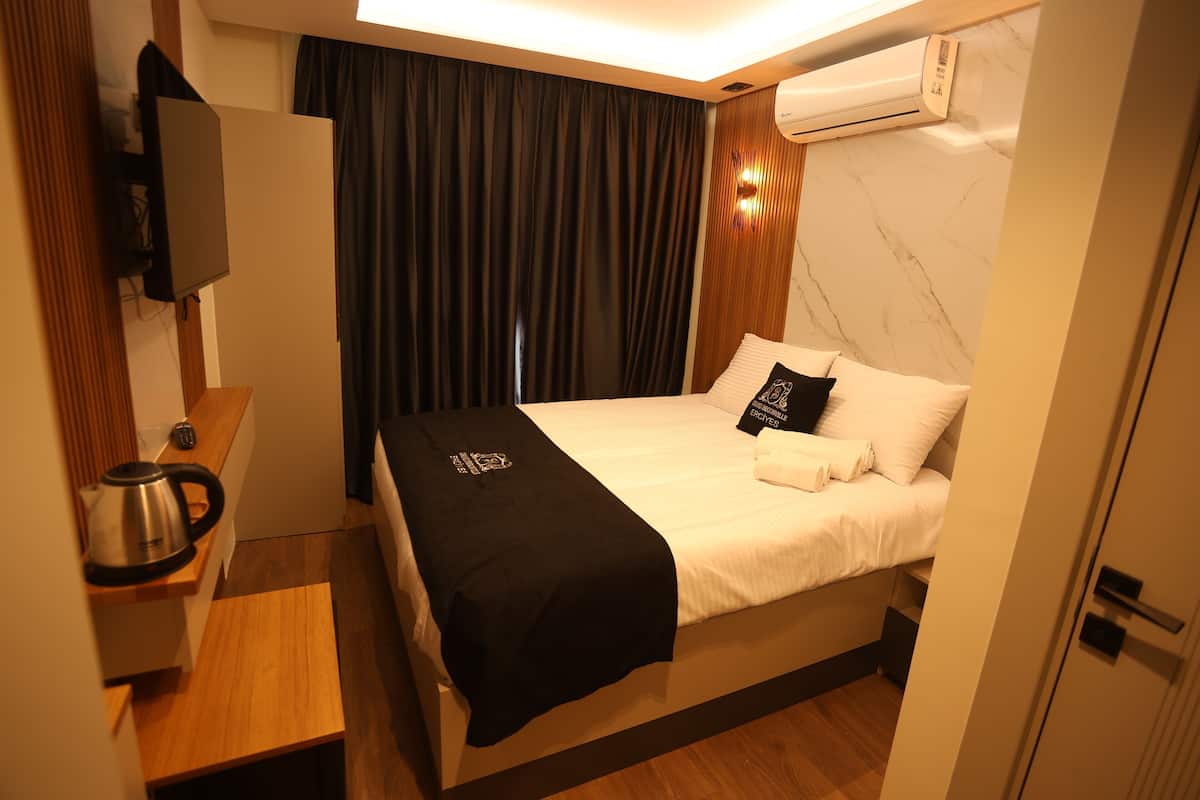 Quarto de hotel com uma cama, uma mesinha de cabeceira com chaleira, um cobertor preto e um ar condicionado fixado na parede.