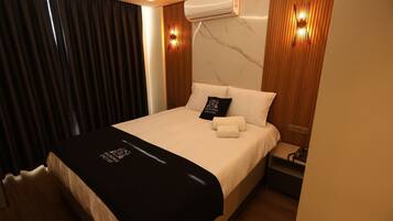 A hotel room with a bed, a nightstand, an air conditioning unit, and a wall-mounted light fixture.