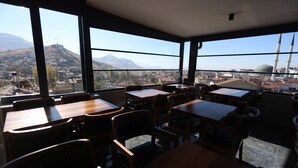 A restaurant with large windows offering a view of a cityscape and mountains. Tables and chairs are arranged for dining. - Grand Begonville Erciyes Hotel (Develi)