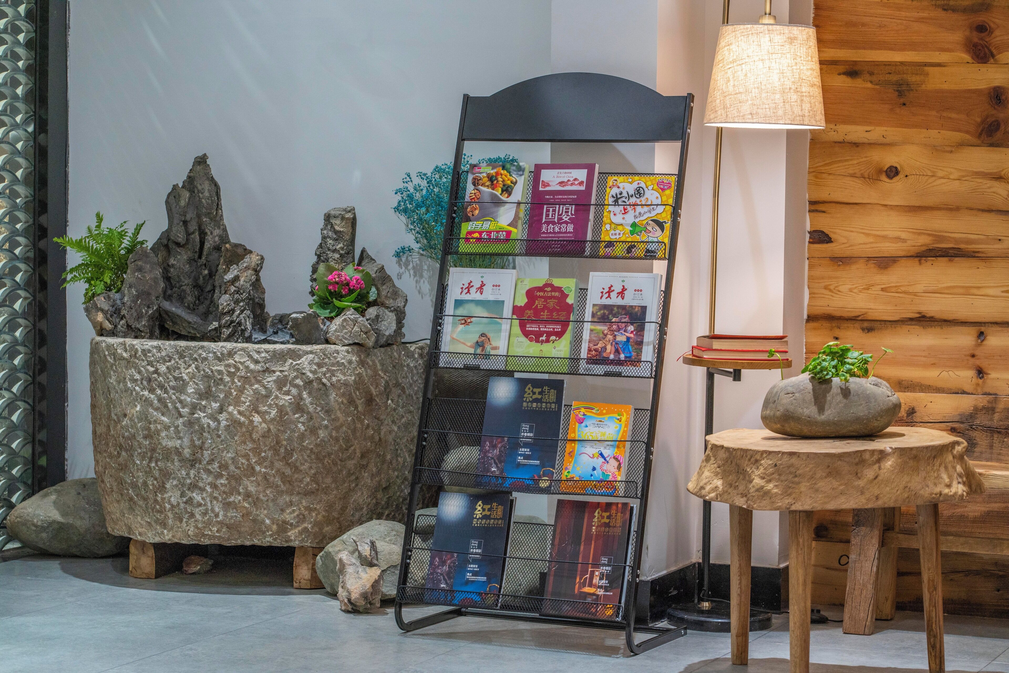 Un rincón acogedor con una jardinera de piedra, un estante metálico para libros y una mesa de madera con una lámpara.
