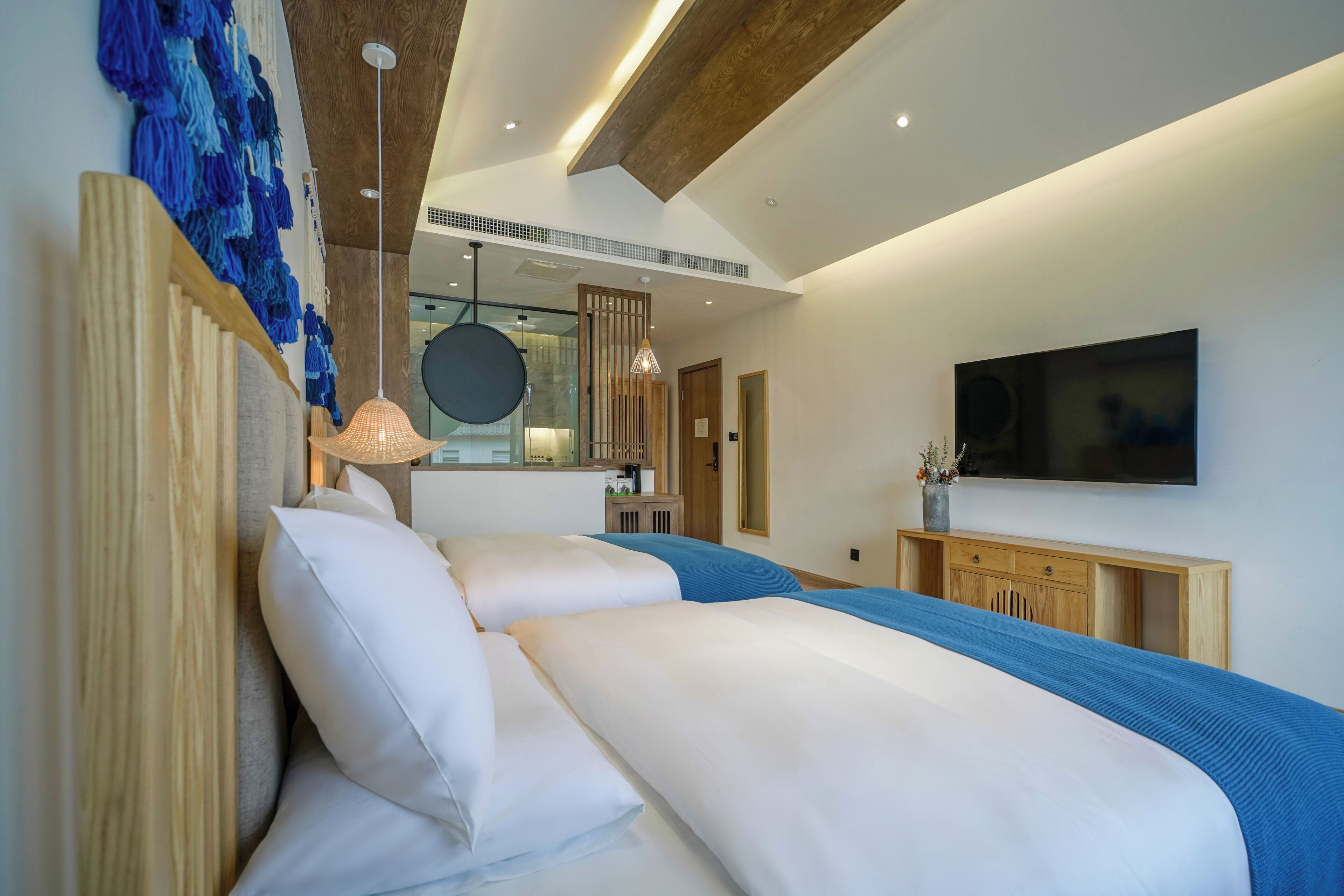 A modern bedroom with a large bed, a TV mounted on the wall, and a kitchen area visible through a glass partition.
