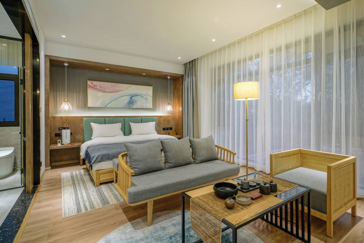 Una habitación de hotel moderna con un sofá de madera, una pequeña mesa con juego de té, una cama con cabecera verde y un ventanal con cortinas translúcidas.