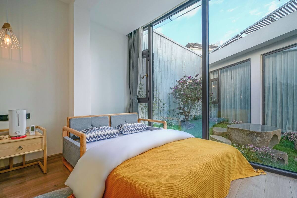 Un dormitorio moderno con un ventanal grande, una cama con una manta amarilla, una mesita de noche con cafetera y vistas a un jardín.