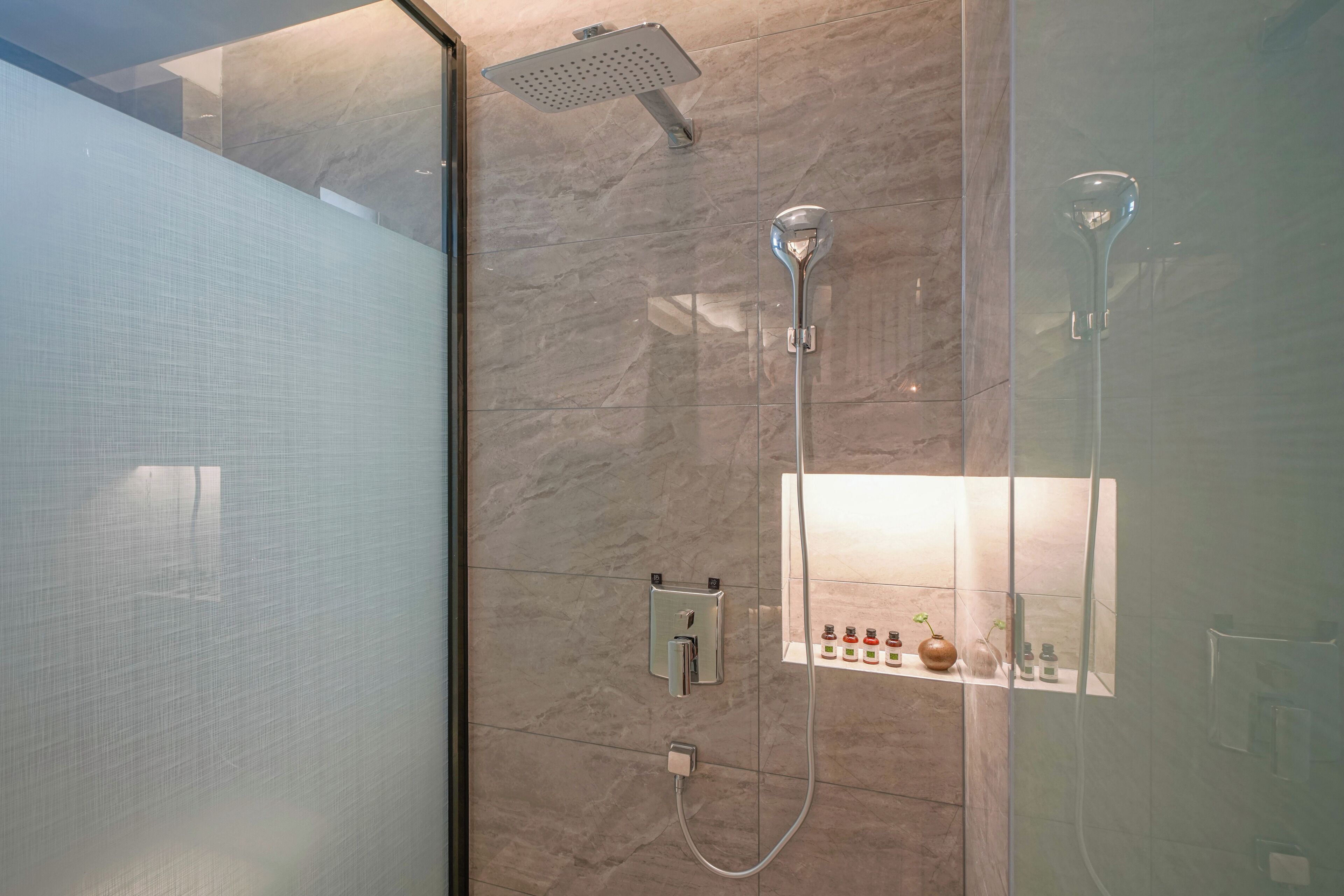 A modern bathroom with a glass shower enclosure, a handheld showerhead, a wall-mounted control panel, and a glass door.