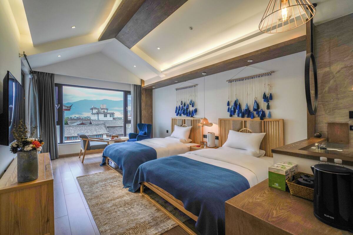 Habitación de hotel con dos camas, un escritorio, una silla, un ventanal con vistas y una lámpara de pared.