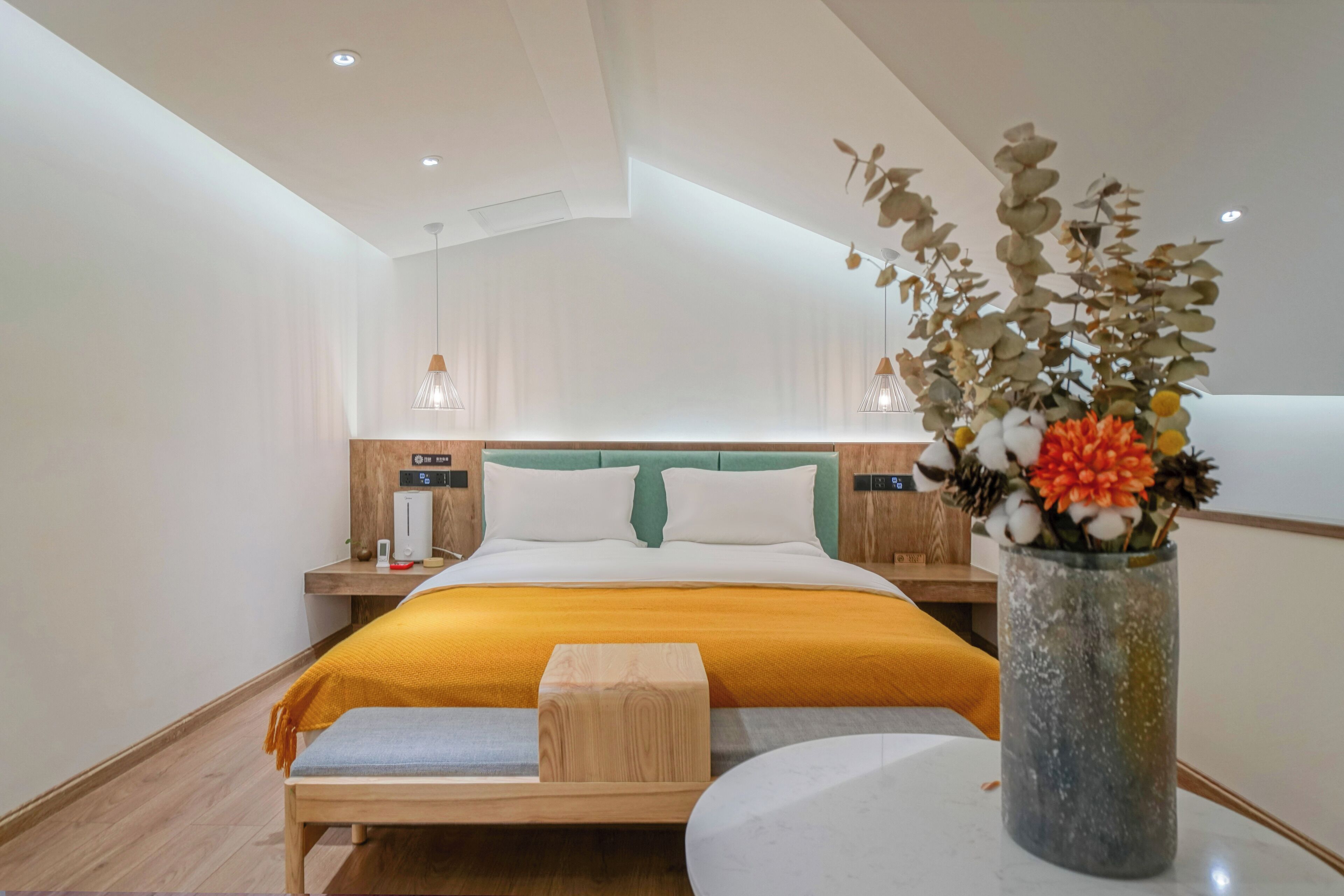 Un dormitorio moderno con una cama, mesitas de noche, una lámpara de noche, un banco de madera y un florero con flores.