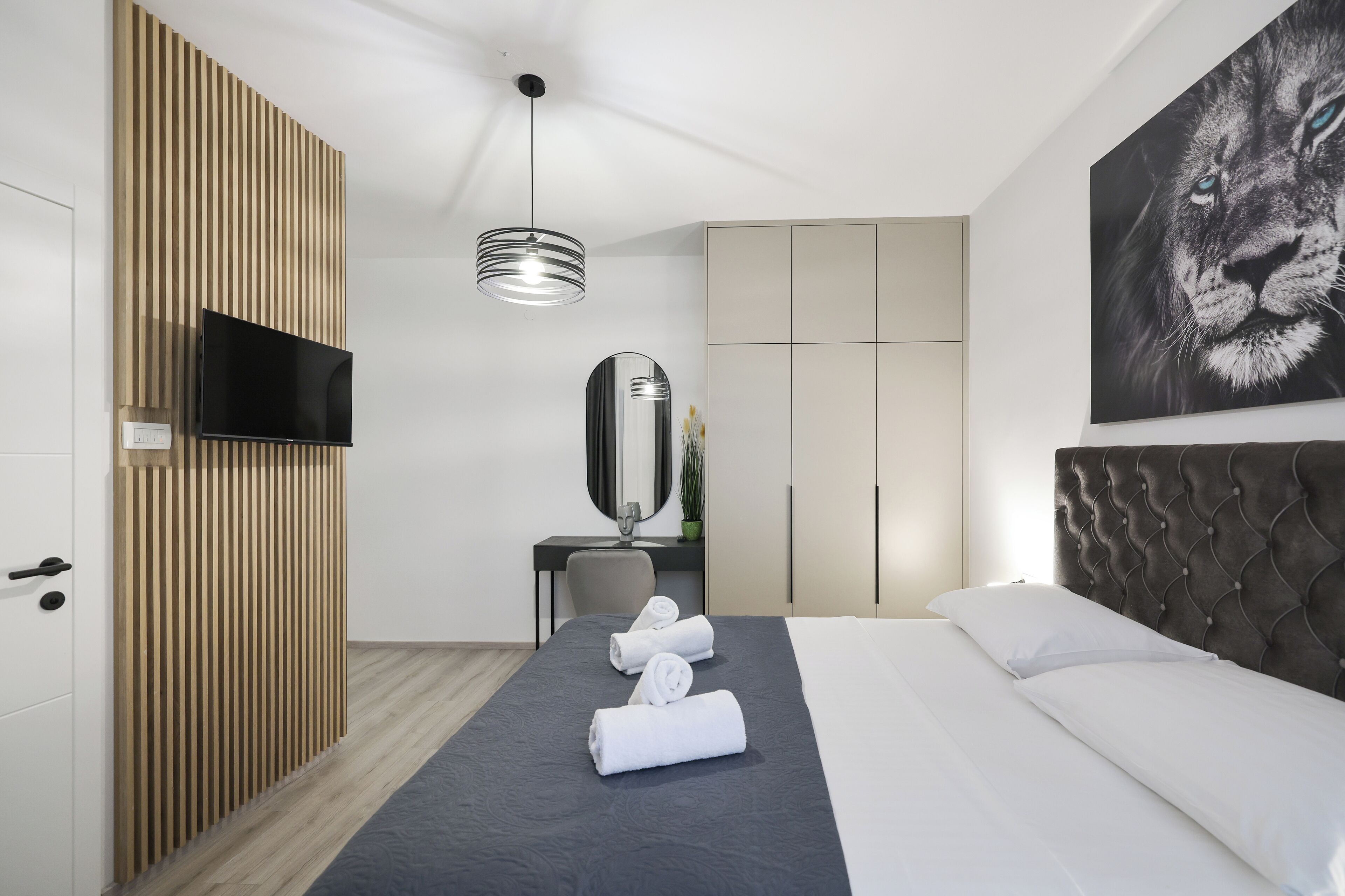 Habitación de hotel moderna con una cama grande, un televisor de pantalla plana, un revestimiento de pared de listones de madera, un espejo amplio y un cuadro con la cara de un león.