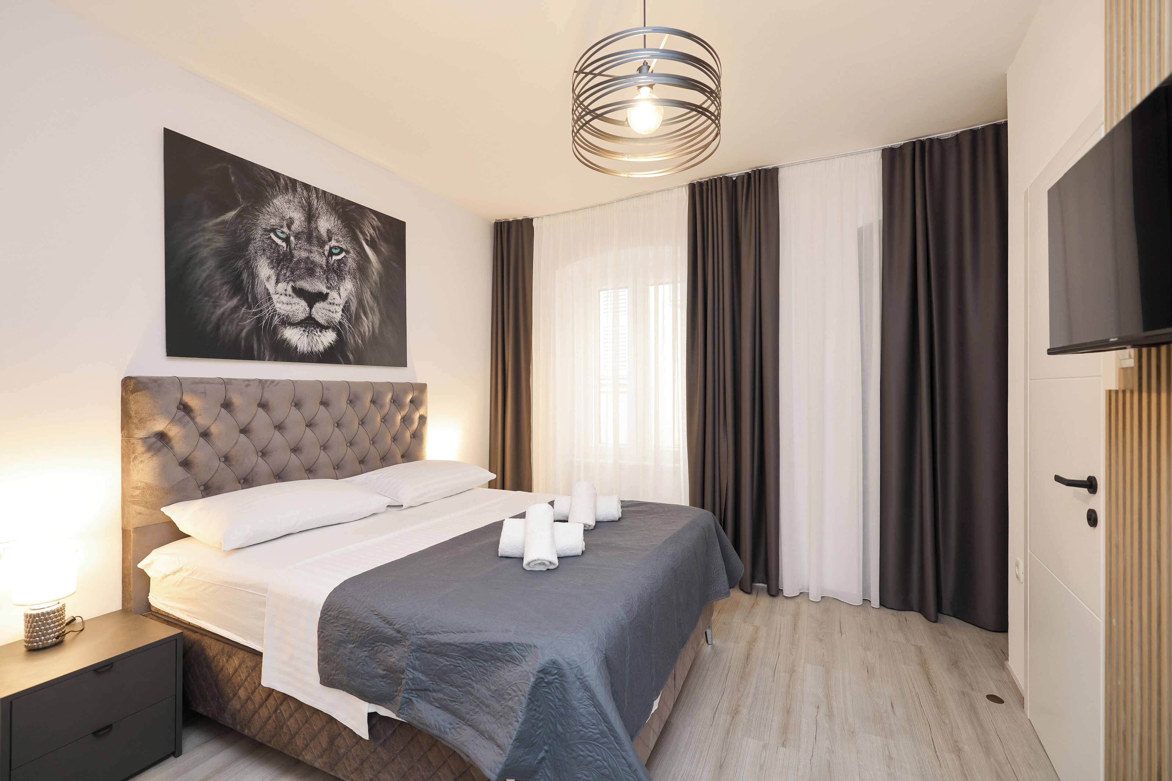 A modern hotel room with a tufted headboard, a large framed lion artwork, a neatly made bed with rolled towels, a flat-screen TV, and a large window with curtains.