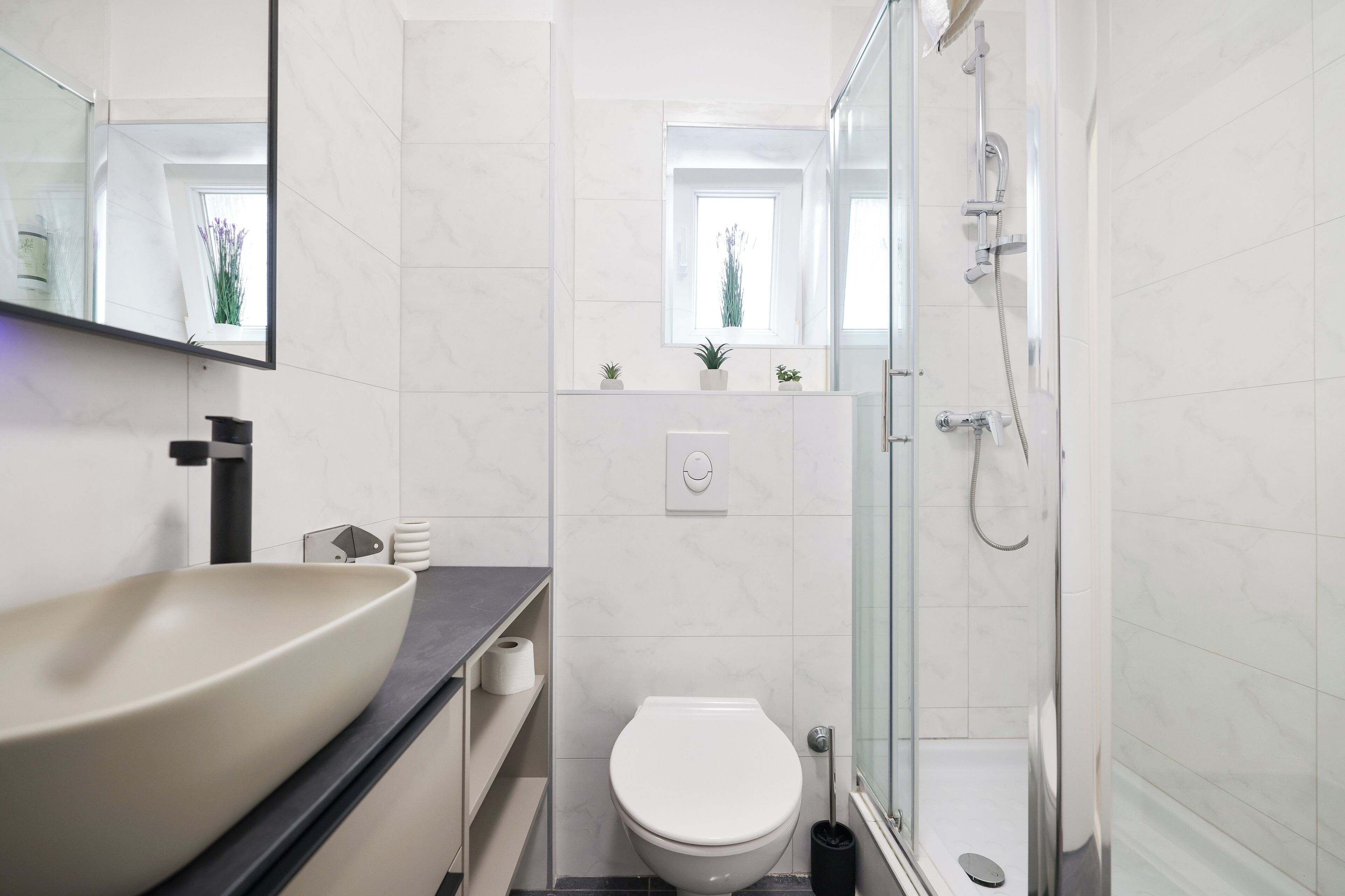 A modern bathroom with a large vessel sink, a toilet, a shower, and a small window with plants.