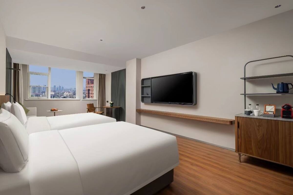 A hotel room with two beds, a TV mounted on the wall, a wooden cabinet, and a window with a city view.