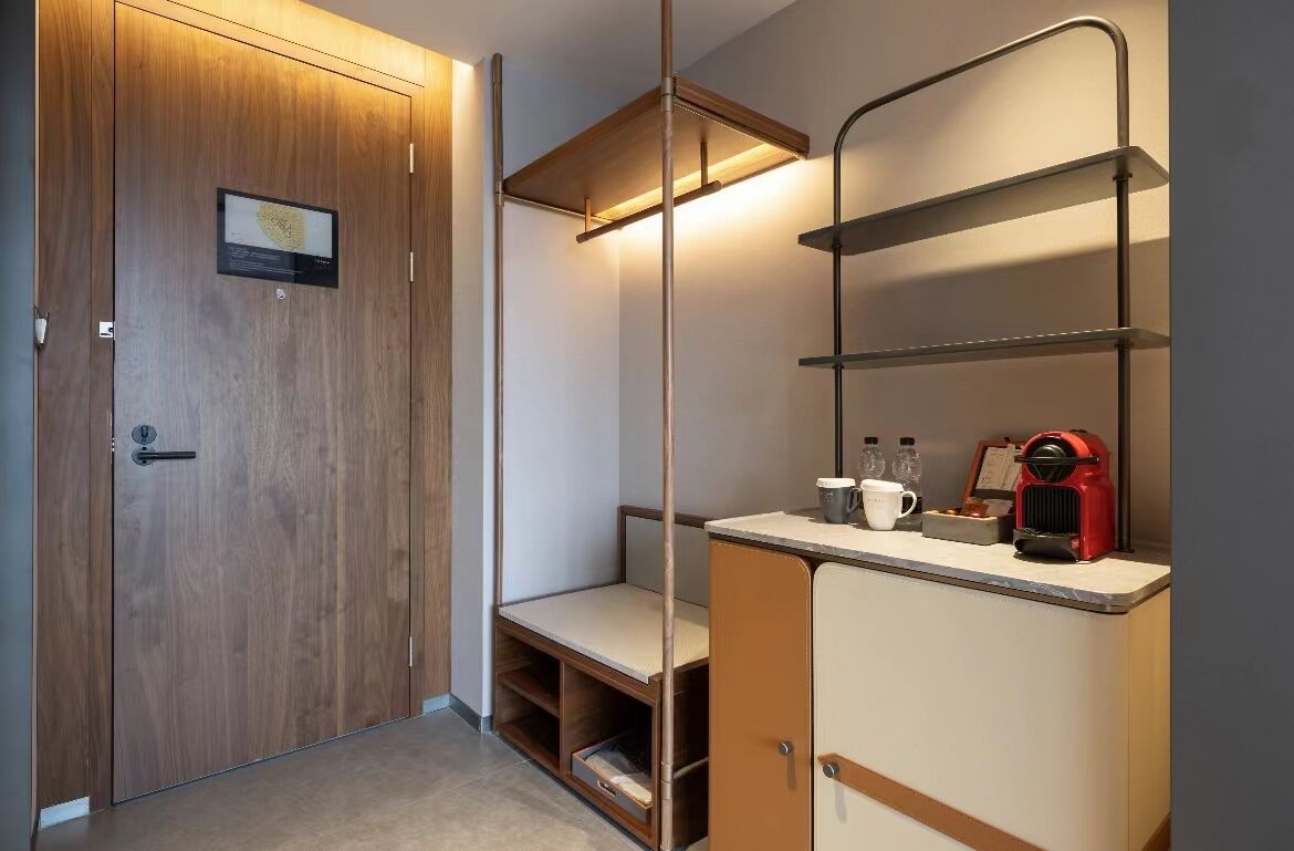 A hotel room with a wooden wardrobe, a bench, a shelf with a coffee maker, and a small table with a cup and a bottle.