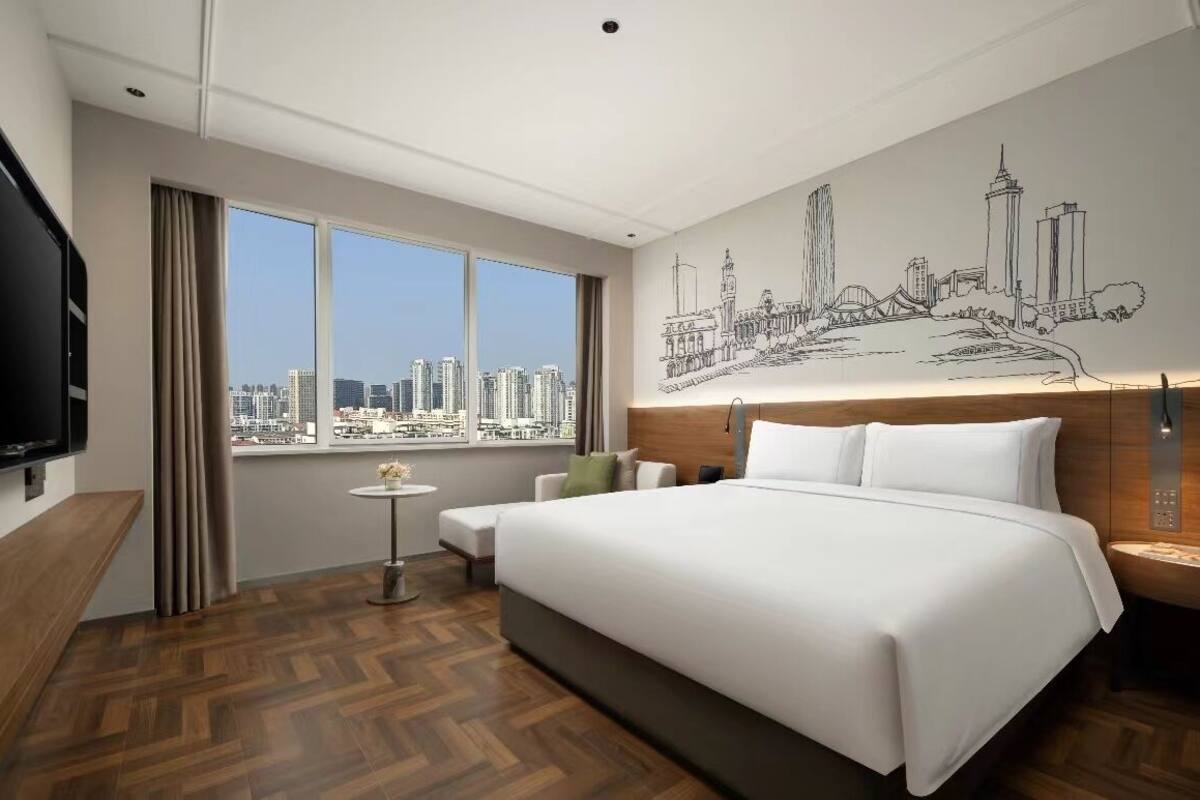 A hotel room with a large bed, a cityscape view, a small table, and a TV on the left wall.