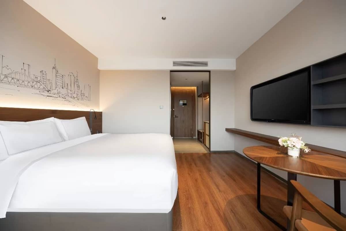 A hotel room with a large bed, a TV mounted on the wall, a wooden table with a vase of flowers, and a cityscape mural on the wall.