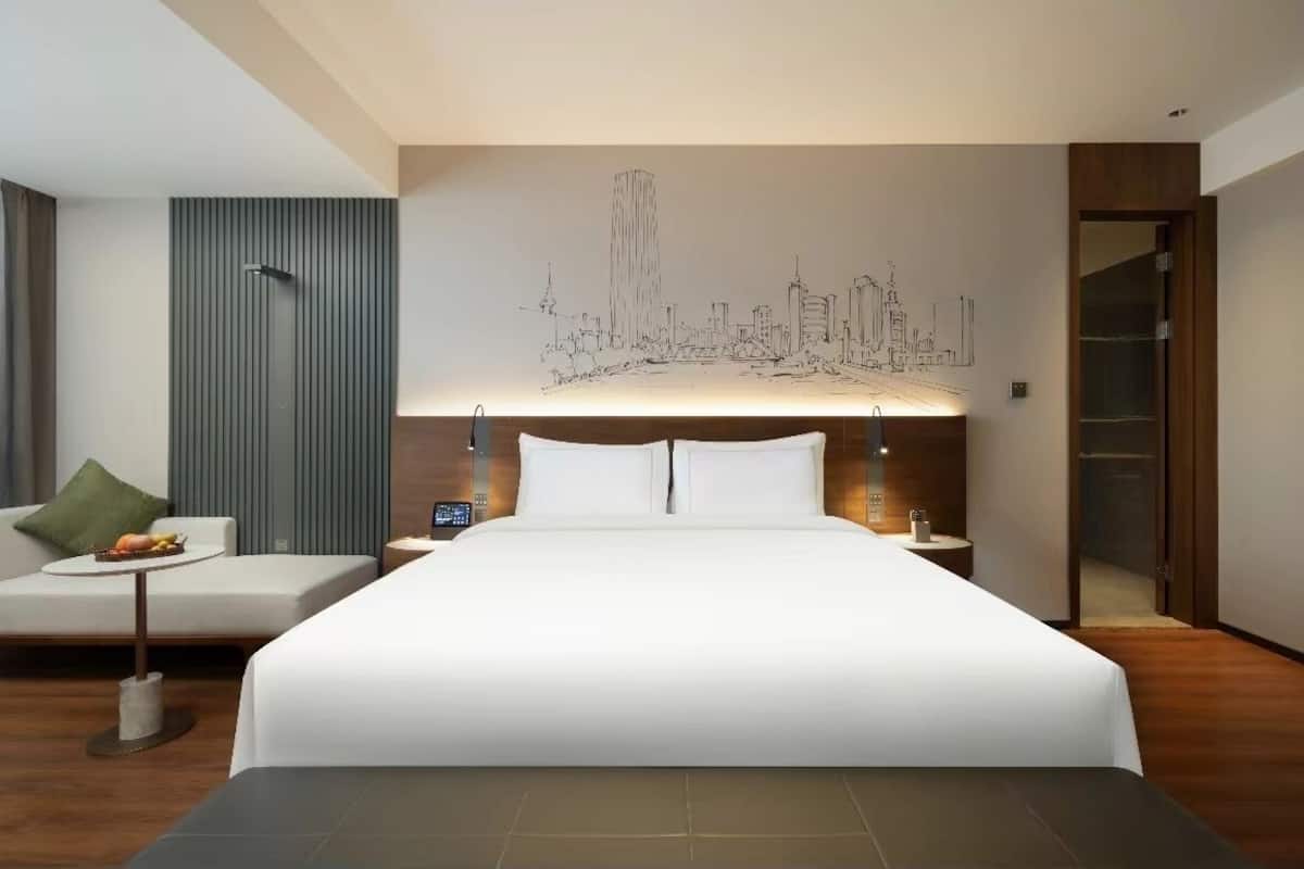 A modern hotel room with a large bed, a bedside table with a lamp, a small side table with a fruit bowl, and a green pillow on a sofa.