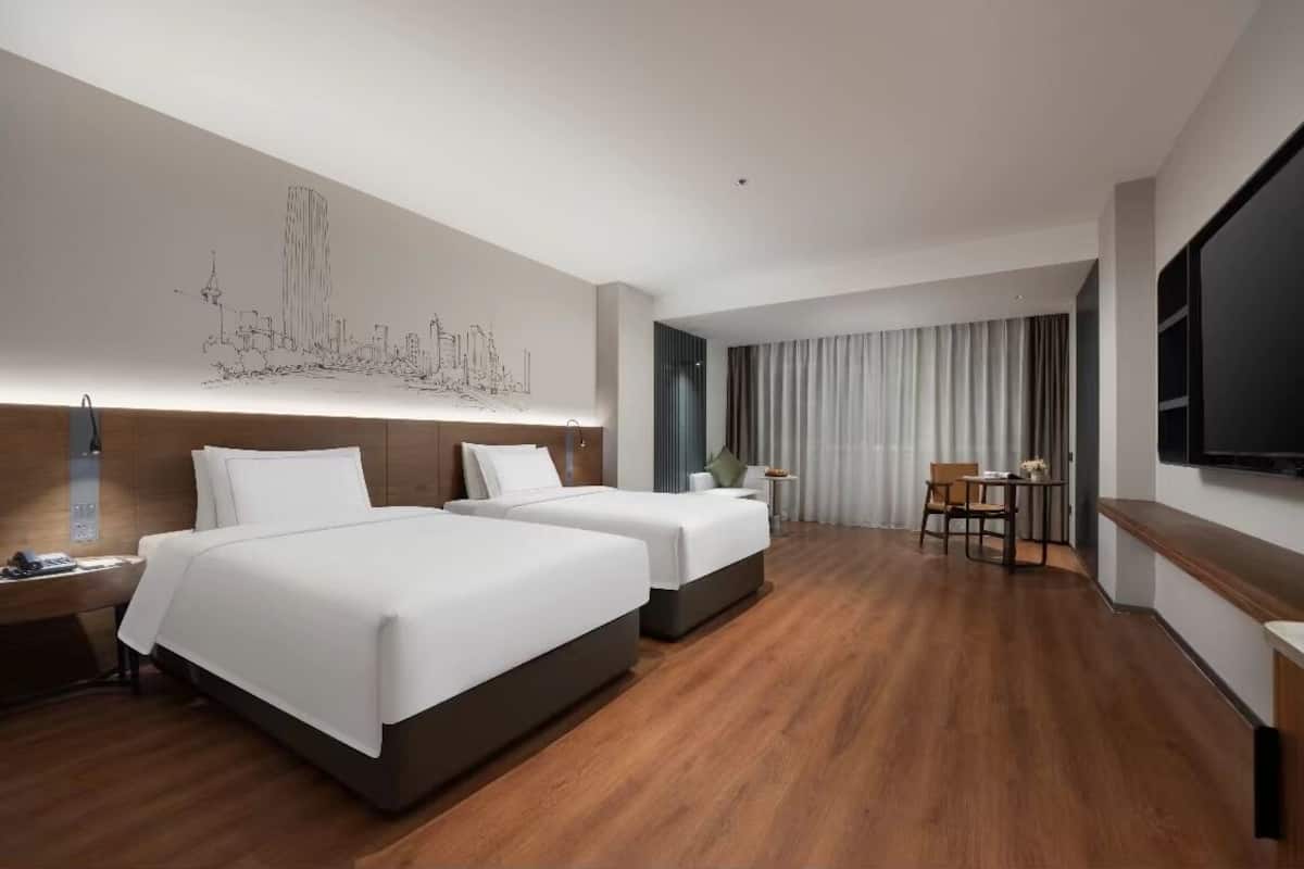 A hotel room with two beds, a desk, a chair, a TV, and a cityscape artwork on the wall.