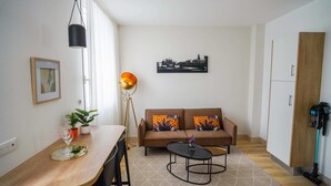 Un soggiorno moderno con un tavolo da pranzo in legno, un divano con cuscini decorati, una lampada da terra e un quadro raffigurante una veduta cittadina.