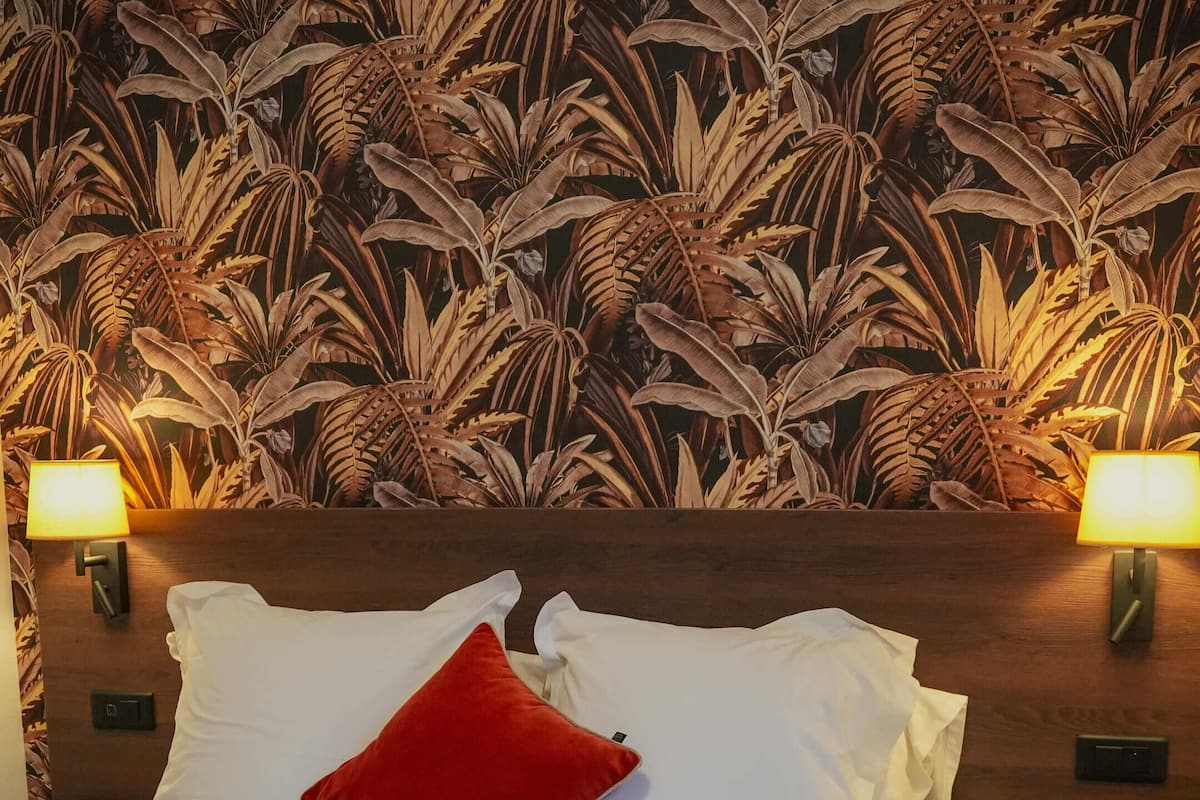A hotel room with a large bed, white bedding, a red throw blanket, and a wall with a tropical leaf pattern. There are two bedside lamps and a wooden headboard.
