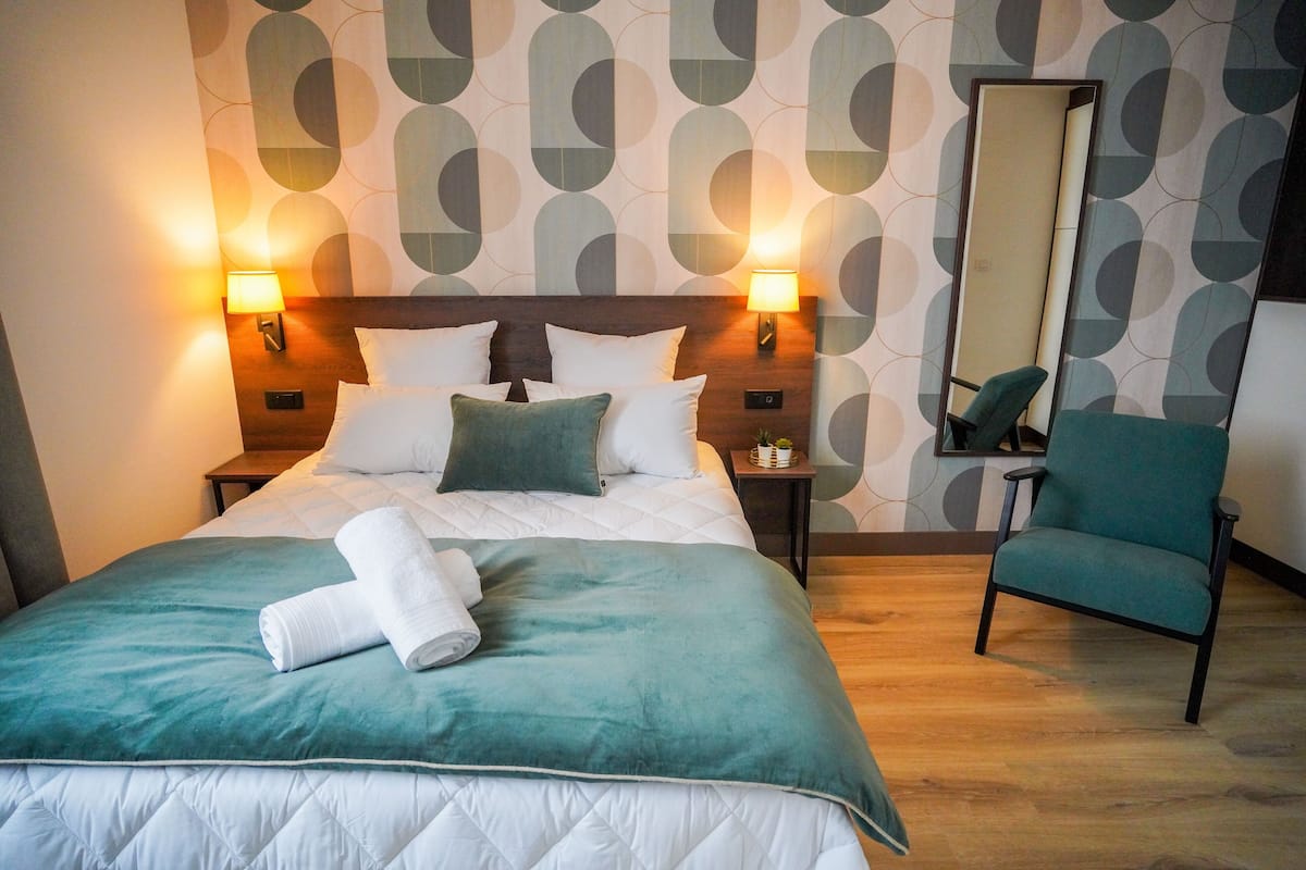 A neatly made bed with a teal bedspread and white pillows, two bedside lamps, a mirror, and a teal armchair in a modern room with patterned wallpaper.