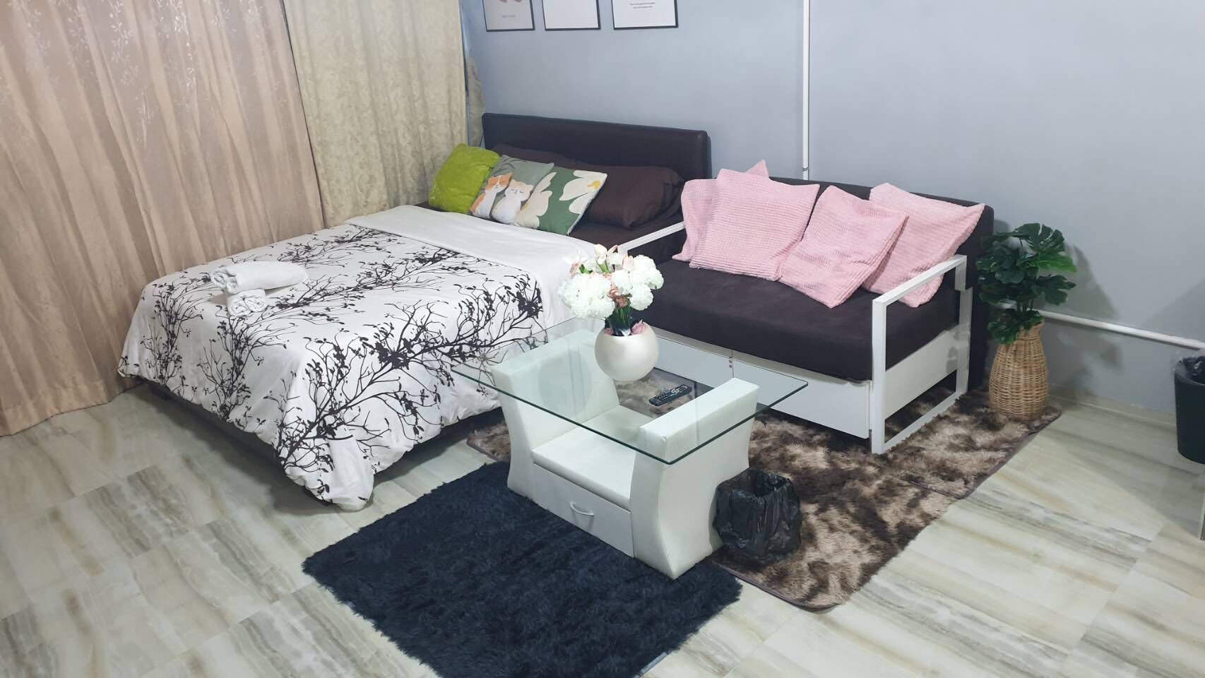 A bedroom with a bed, a white sofa, a glass coffee table, a black rug, and a potted plant.