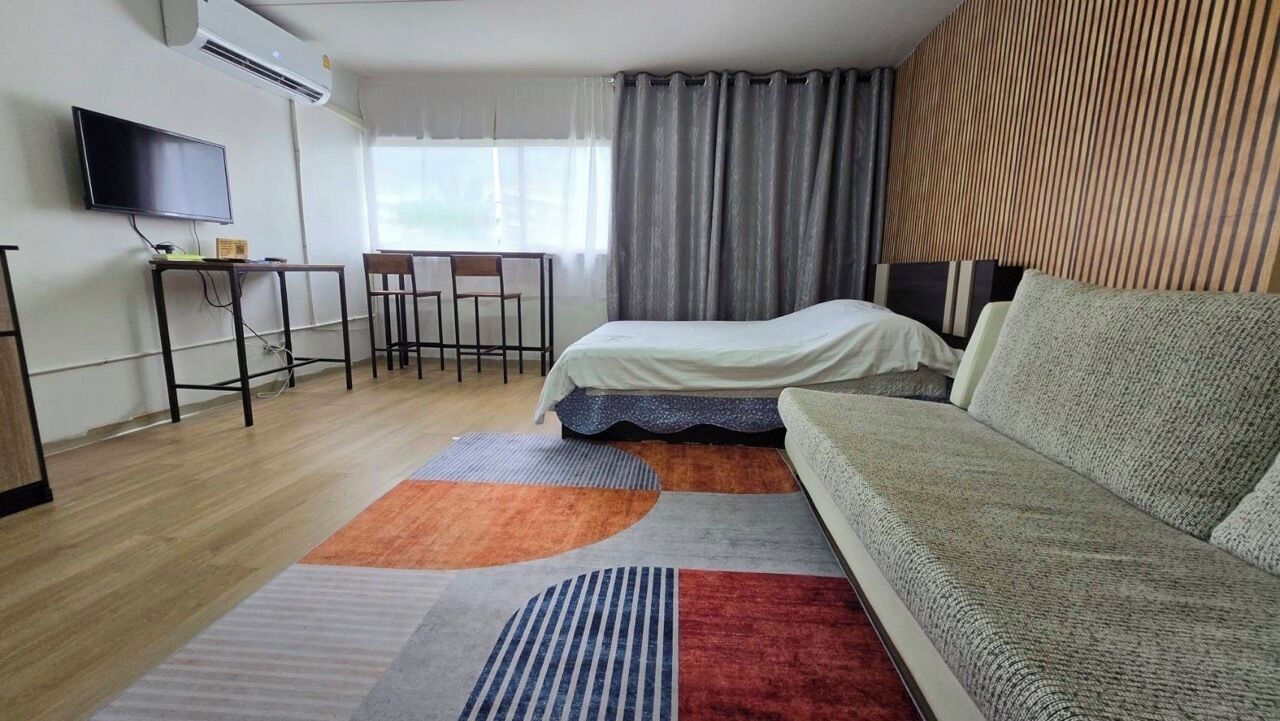 A modern hotel room with a bed, a sofa, a desk with a chair, a flat-screen TV, and a large window with curtains.