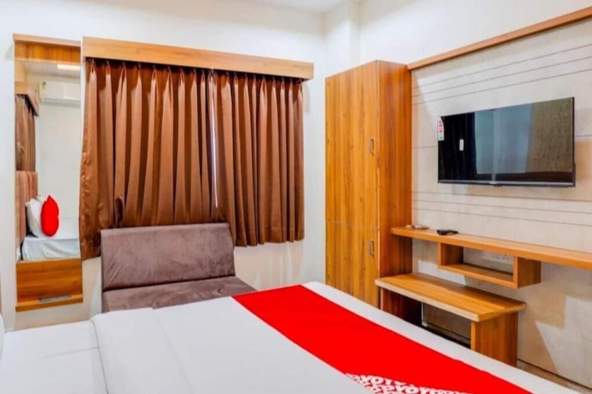 A hotel room with a bed, a TV mounted on the wall, wooden furniture, and a window with curtains.