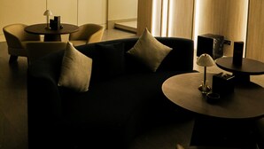 Een moderne hotelkamer met een donkergroene bank, een ronde salontafel en een klein bijzettafeltje met een lamp. Er zijn twee stoelen, een groot raam met verticale jaloezieën en een wandgemonteerde airconditioning.