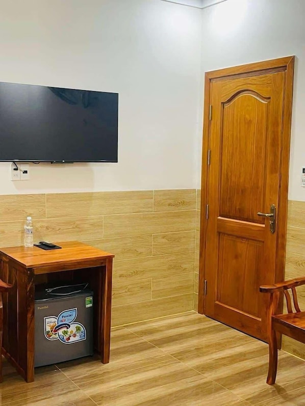 A room with a wooden door, a television mounted on the wall, a small wooden table with a water bottle and a mobile phone, and a trash bin with a sticker. - Hotel Hoàng Anh (Dau Giay)
