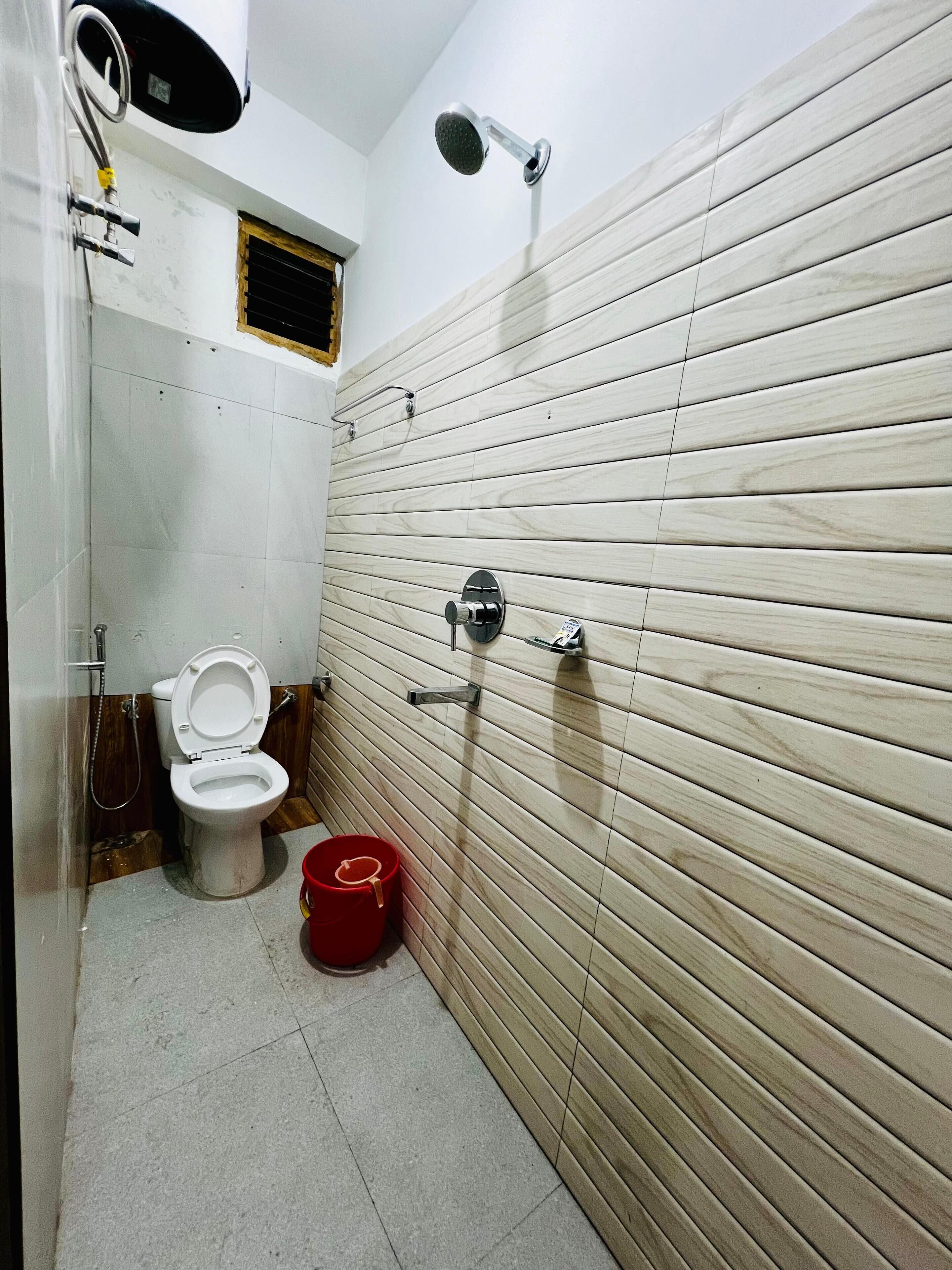 A bathroom with a toilet, a shower, and a red bucket on the floor.