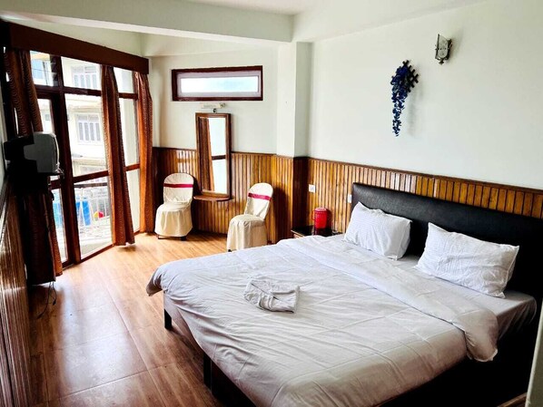 A hotel room with a large bed, a wooden headboard, a window with curtains, a chair, a small table, and a mirror. - HOTEL TASHI GYAL TSEN (Gyalshing)