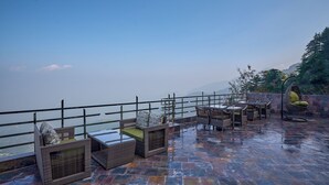 Un espace extérieur aménagé avec des meubles en osier et une table en verre, offrant une vue sur un plan d’eau et des montagnes.