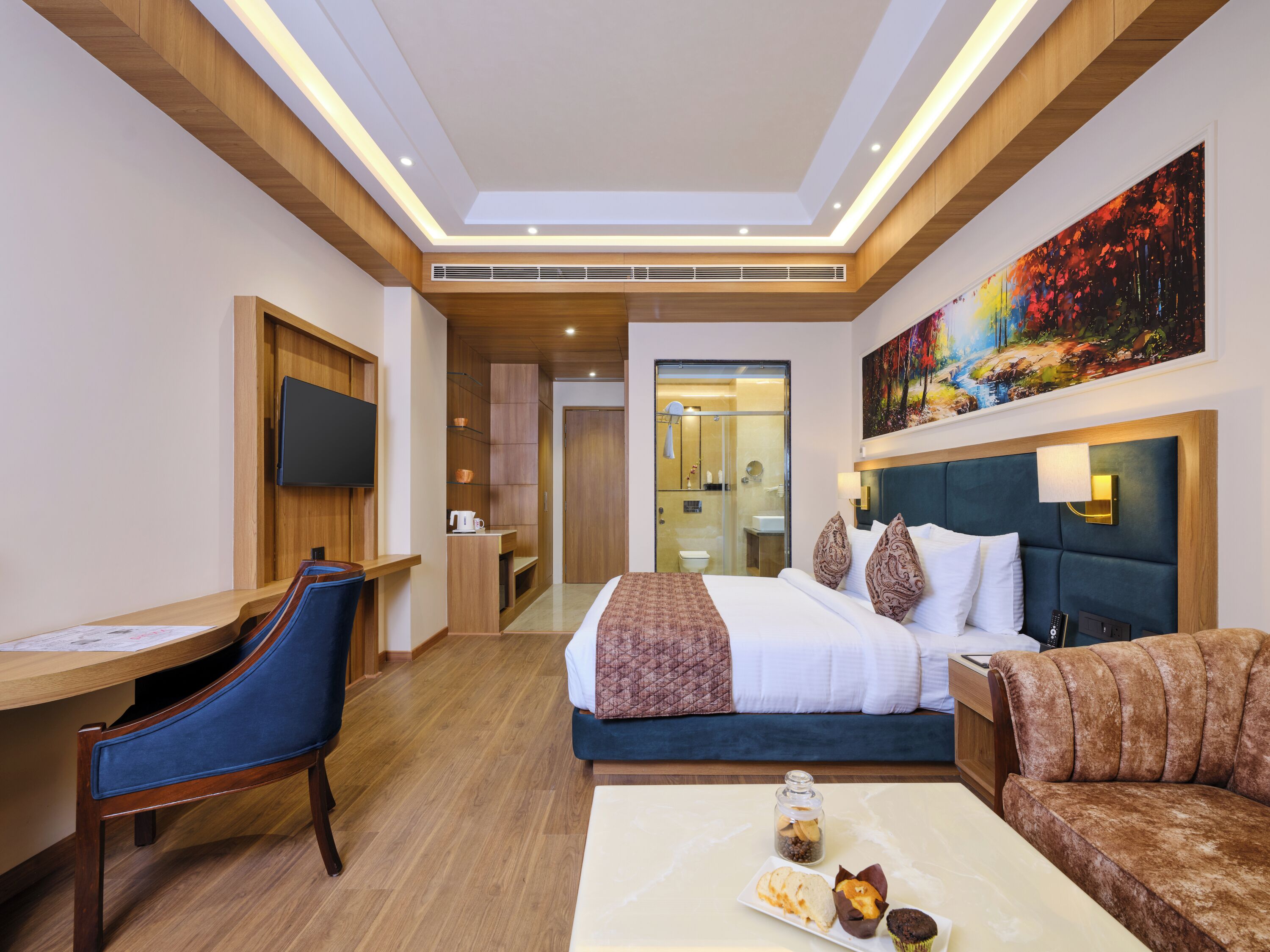 Une chambre d’hôtel avec un lit, un bureau, une chaise, un canapé, une table basse avec des pâtisseries et un tableau au mur.