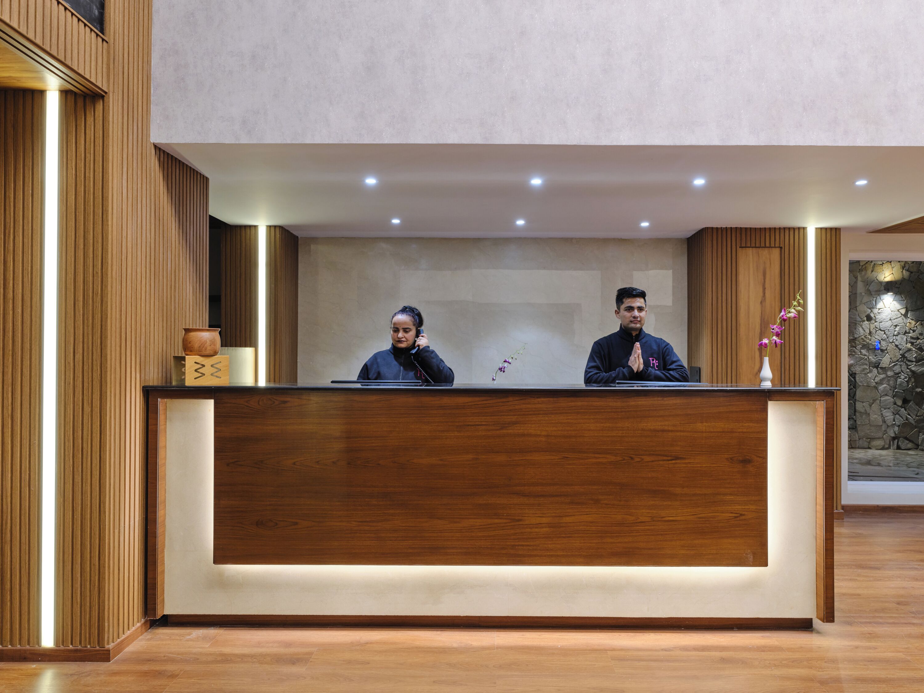 A reception desk with two individuals seated behind it, a vase with flowers, and a stone wall in the background.