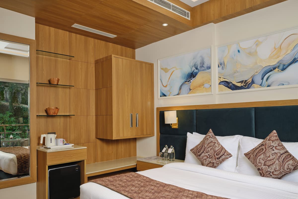 A modern hotel room with a large bed, wooden wardrobe, bedside table with a kettle, and abstract wall art.