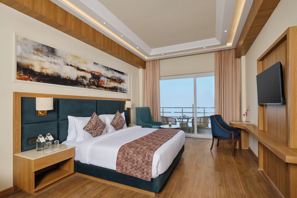 A hotel room with a large bed, a desk with a lamp, a TV, and a balcony with a table and chairs.